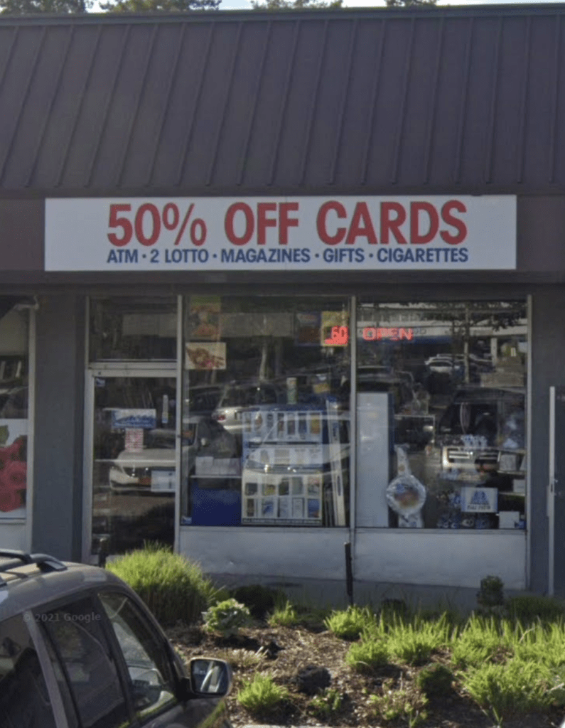 Storefront with a sign advertising "50% OFF CARDS" and other items like "ATM," "2 LOTTO," "MAGAZINES," "GIFTS," and "CIGARETTES."